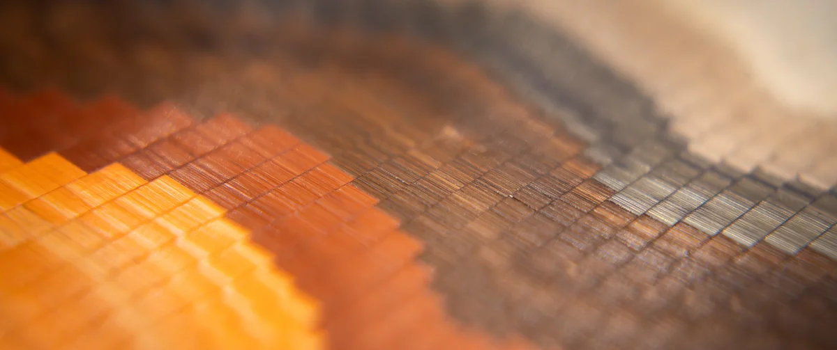 Macro view of straw marquetry laid in stepped, pixel-like blocks, shading from bright orange through copper and brown to grey and cream