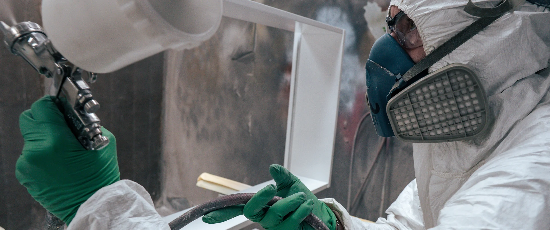 A finisher in a white suit, respirator and green gloves spraying lacquer onto a white furniture frame in the spray booth