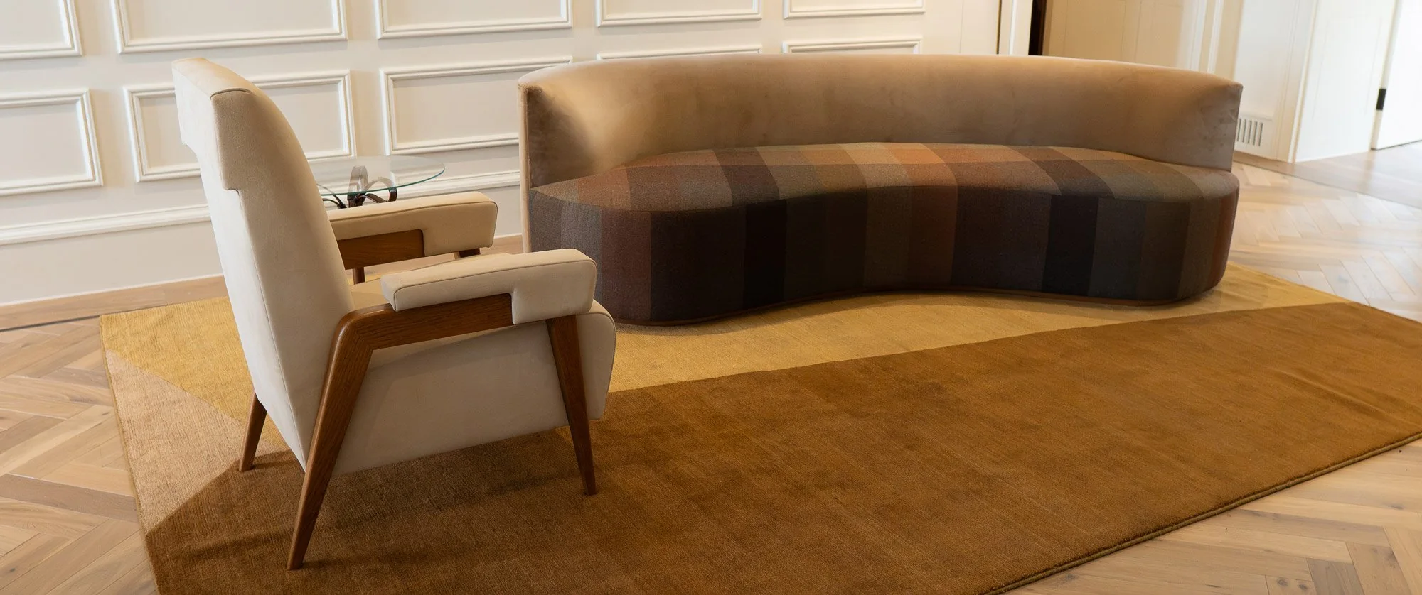 Curved sofa in striped brown and taupe upholstery beside a cream armchair with oak arms in a panelled room