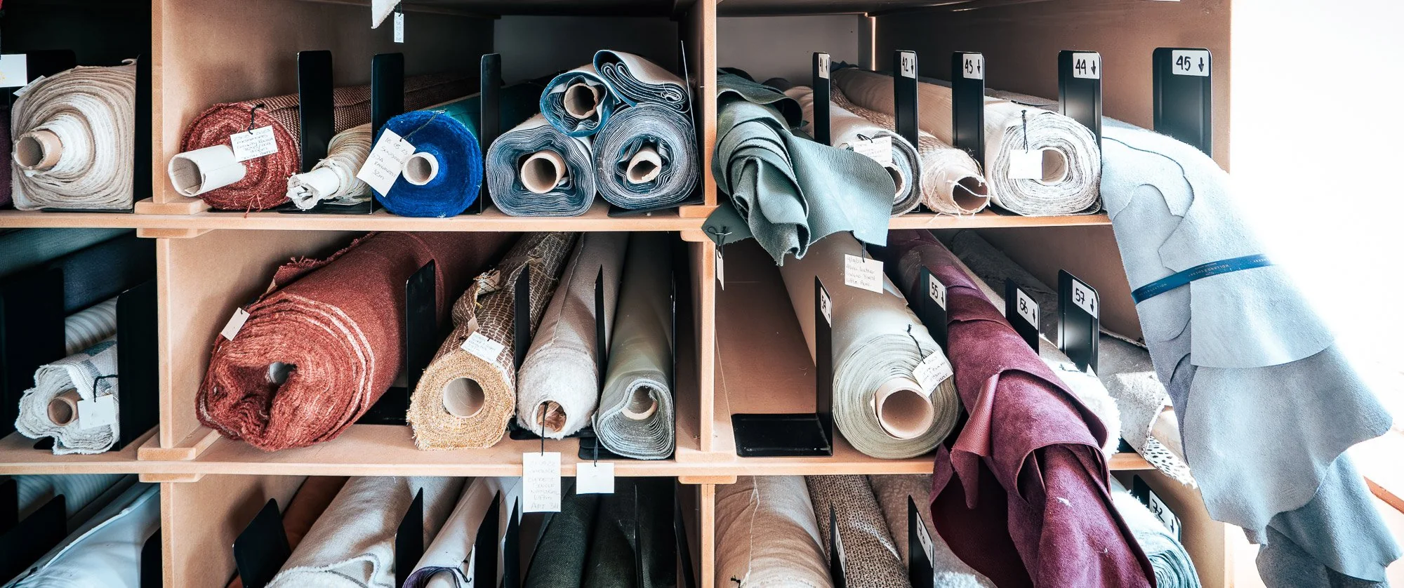 Shelves of rolled upholstery fabrics in blue, rose and cream in the workshop store