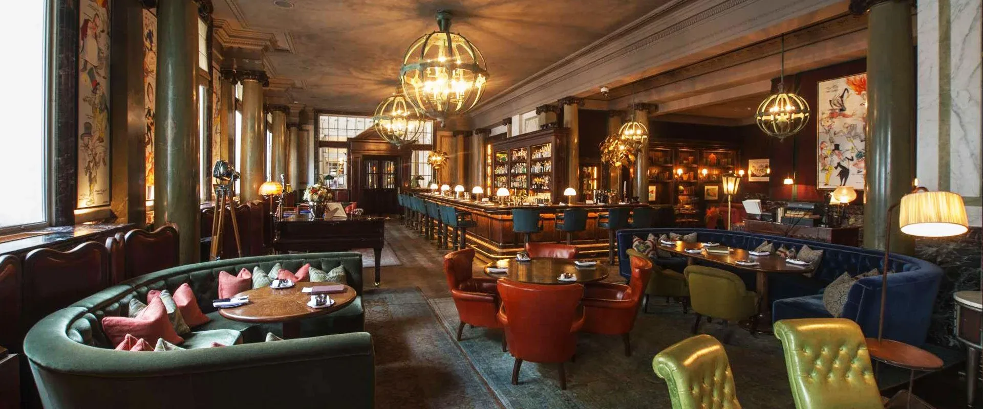 Bar et salon d’hôtel avec des suspensions lanternes sphériques vert-de-gris au-dessus d’un long comptoir, des banquettes vertes et des fauteuils en cuir