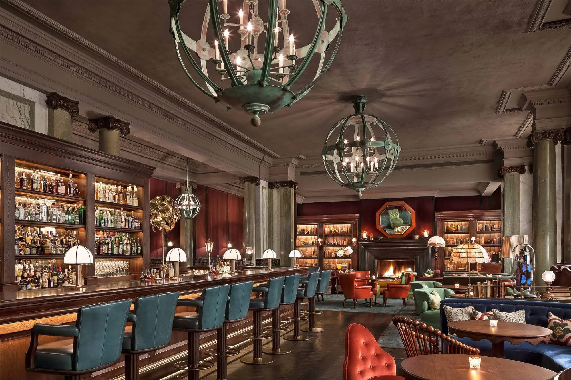 Hotel bar and library lounge with large verdigris globe chandeliers above a long bar, teal leather stools, lit bookshelves and a fireplace
