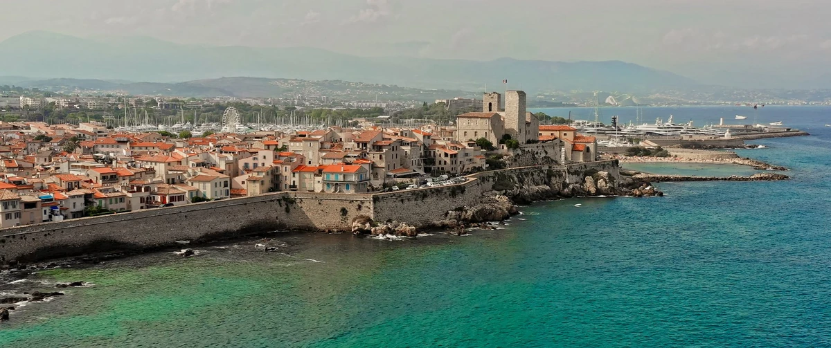 Aerial view of Antibes on the French Riviera, with the walled old town, terracotta roofs, the fort tower and the marina beyond clear turquoise water.