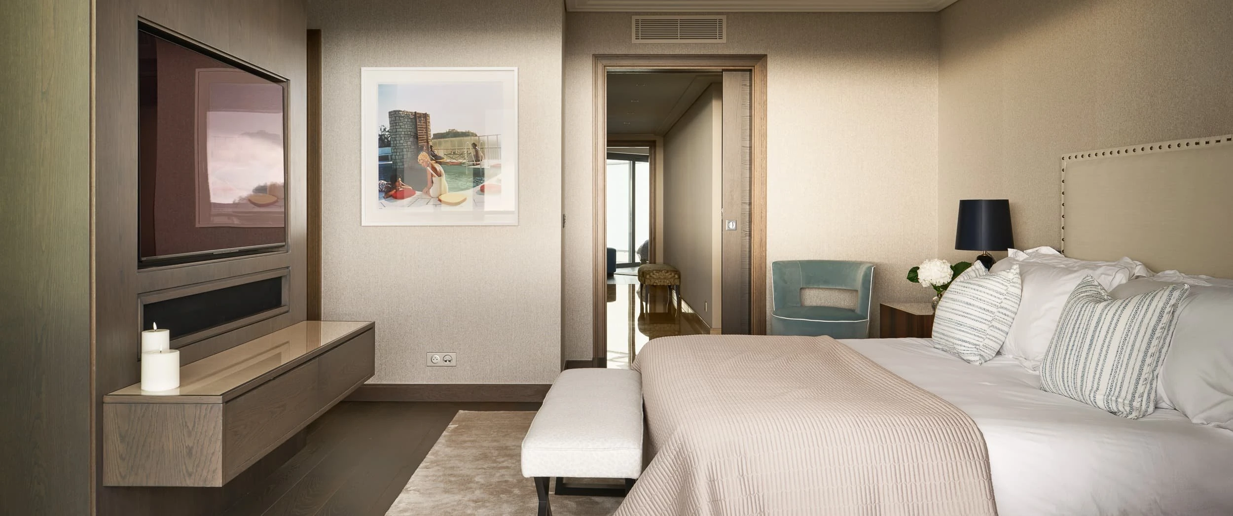 Principal bedroom of the Antibes penthouse, with a studded upholstered headboard, quilted bedcover, framed photograph and an upholstered bench at the foot of the bed.