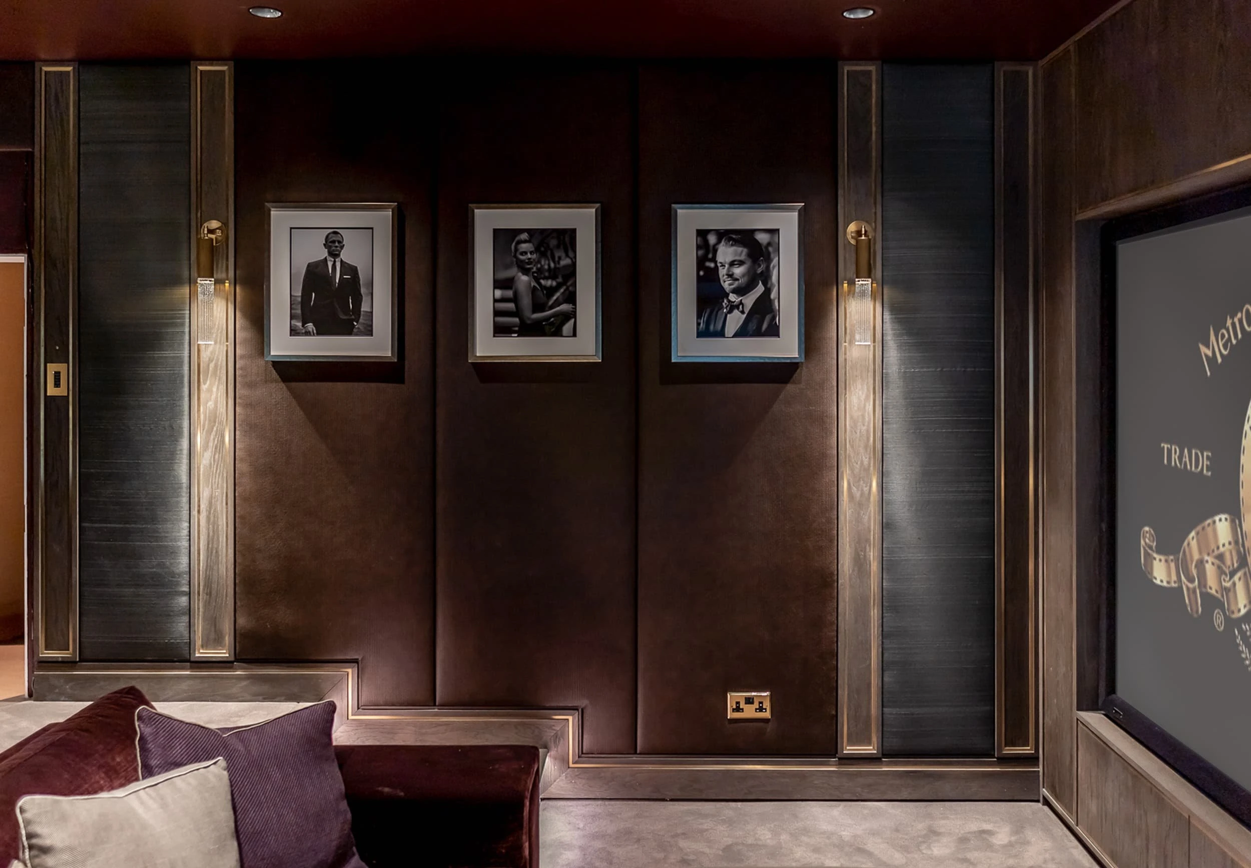 Wide view of the Country Home Cinema showing leather panels with brass inlays, framed portraits, brass wall lights and the edge of the projection screen displaying a studio ident.