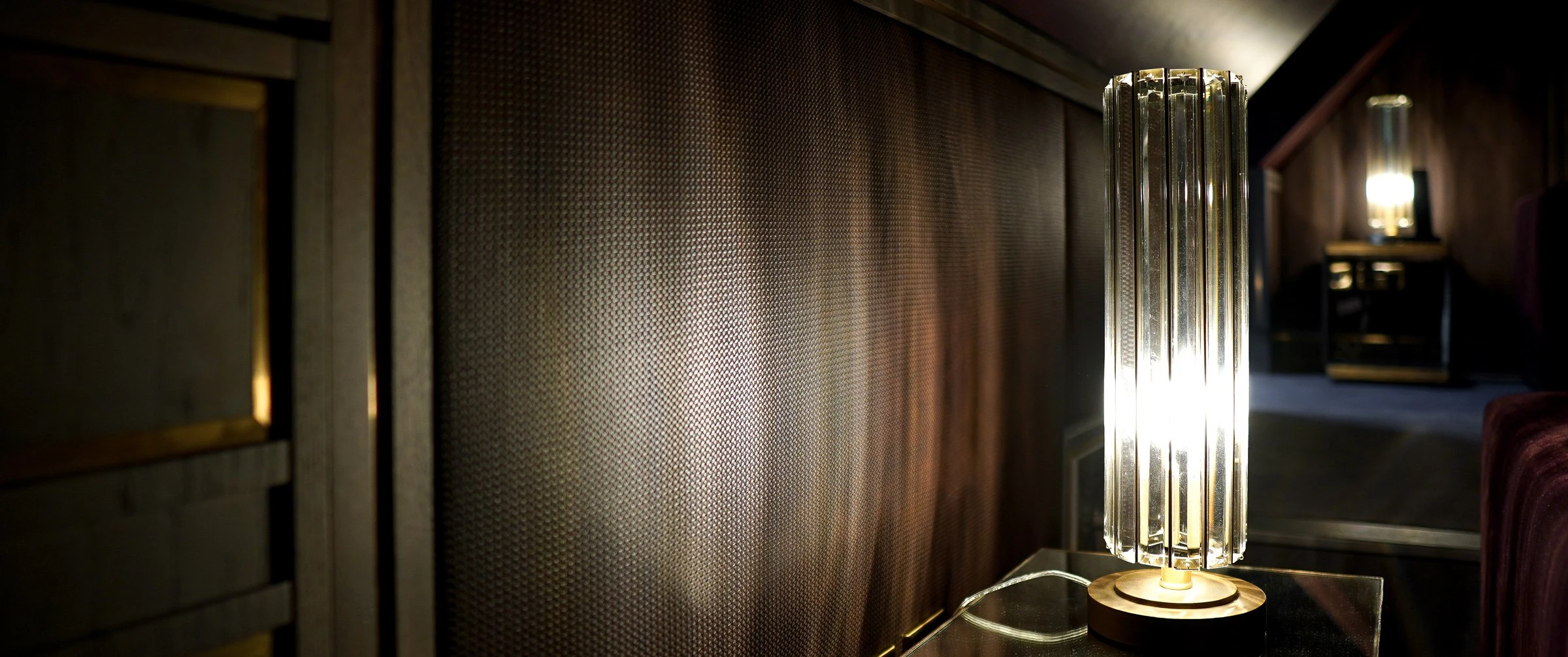 Fluted crystal table lamp lit on a dark side table against woven leather panelling in the Country Home Cinema, with a second lamp glowing in the background.