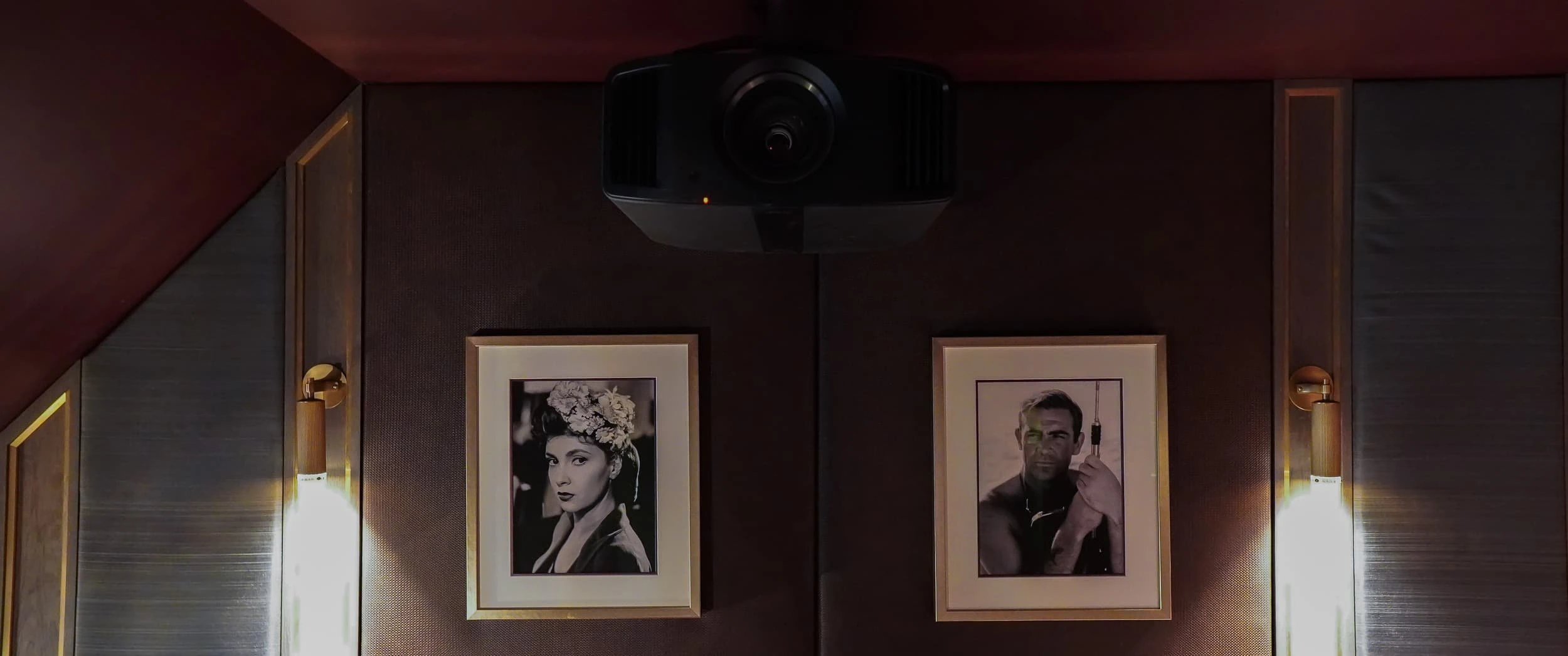 Ceiling-mounted projector fixed to the burgundy ceiling above two framed black-and-white portraits and brass wall lights in the Country Home Cinema.