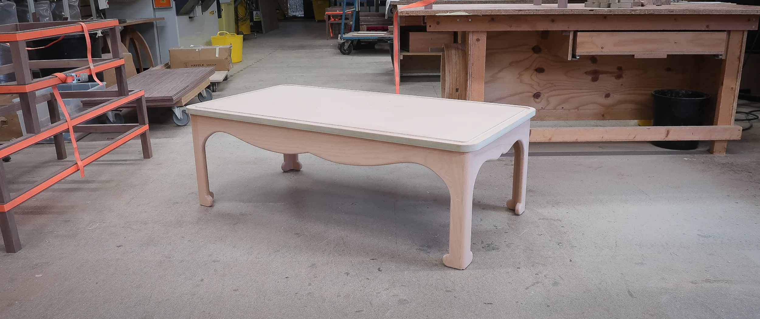 A bespoke low table with a shaped, scalloped apron and carved bracket feet, in bare timber before finishing, in the workshop.