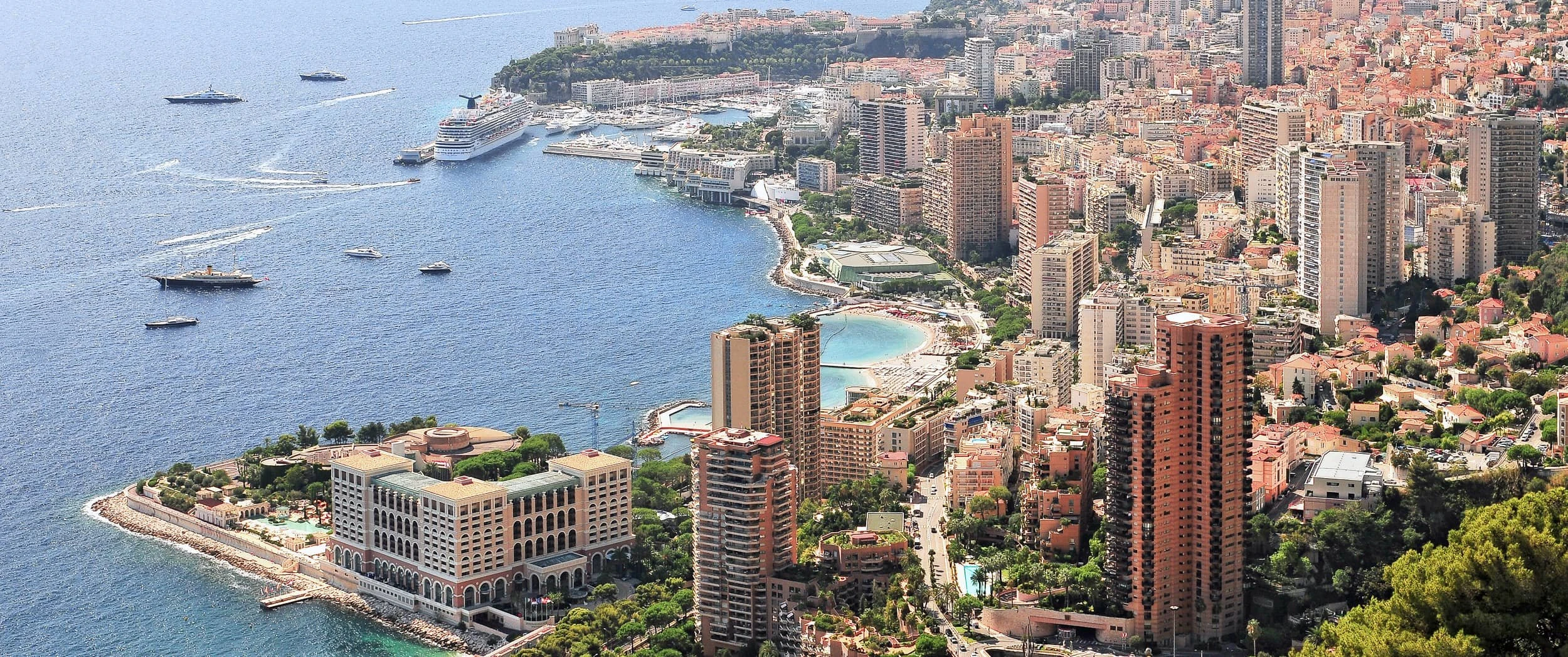 Daytime aerial view of Monaco's harbour, towers and beach seen from the hills near Roquebrune-Cap-Martin, with yachts moored offshore.