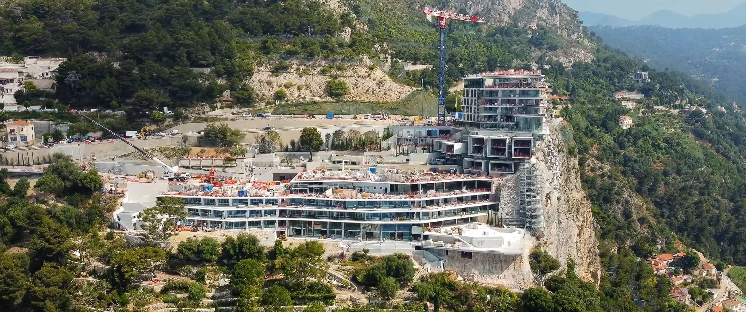 Construction view of the hotel's stepped concrete and glass floors at Roquebrune-Cap-Martin, with cranes, scaffolding and site traffic on the road above.