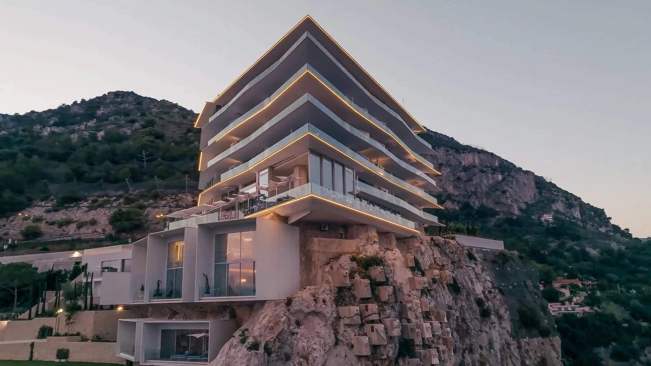 Twilight view of the hotel's illuminated slab edges and terrace level above the cliff face, with the hillside and anchor pads visible below.