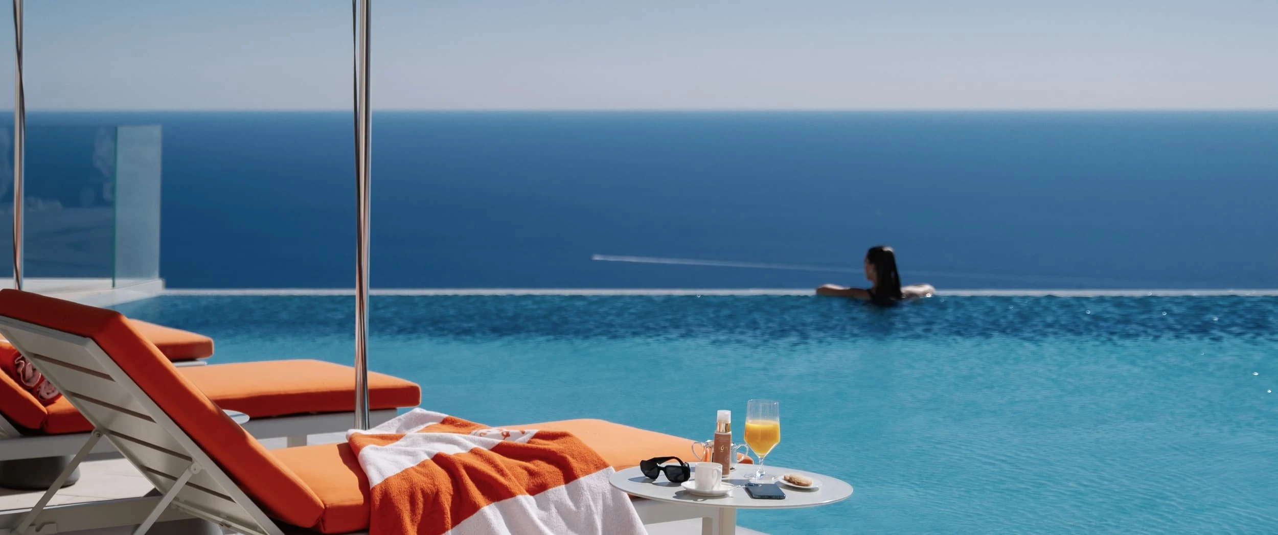 Orange-cushioned loungers and a poolside table with juice and coffee beside the infinity pool at The Maybourne Riviera, a swimmer at the far edge above the sea.