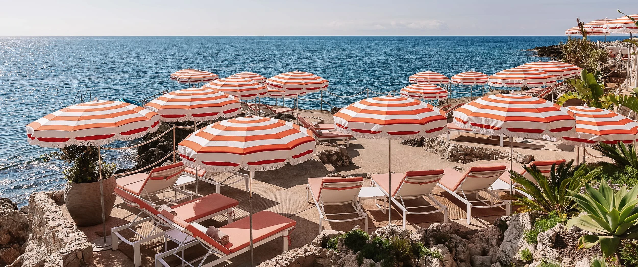 Orange-and-white striped fringed parasols and matching sun loungers on a stone beach terrace beside the Mediterranean at Maybourne La Plage.