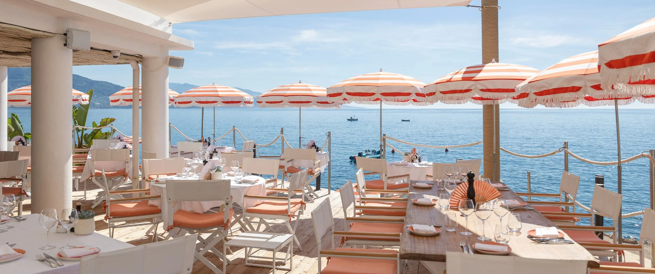 Timber deck restaurant at the water's edge with white folding chairs, orange cushions, striped fringed parasols and rope barriers over the sea.
