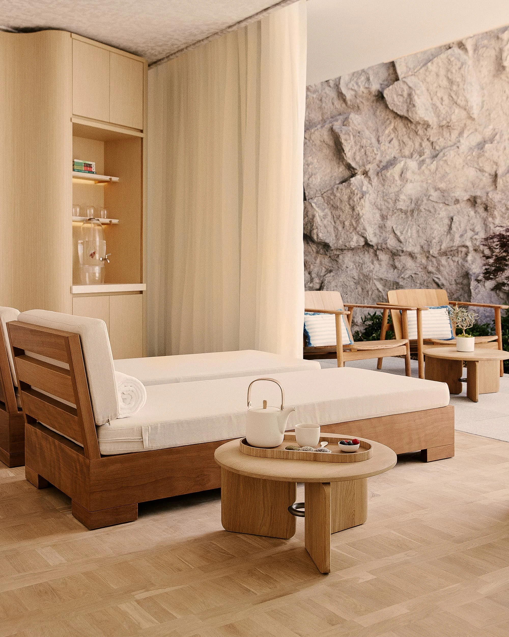 Timber-framed daybeds with pale cushions and a round oak table set with a teapot in the spa relaxation area, a rough rock wall beyond the glazing.
