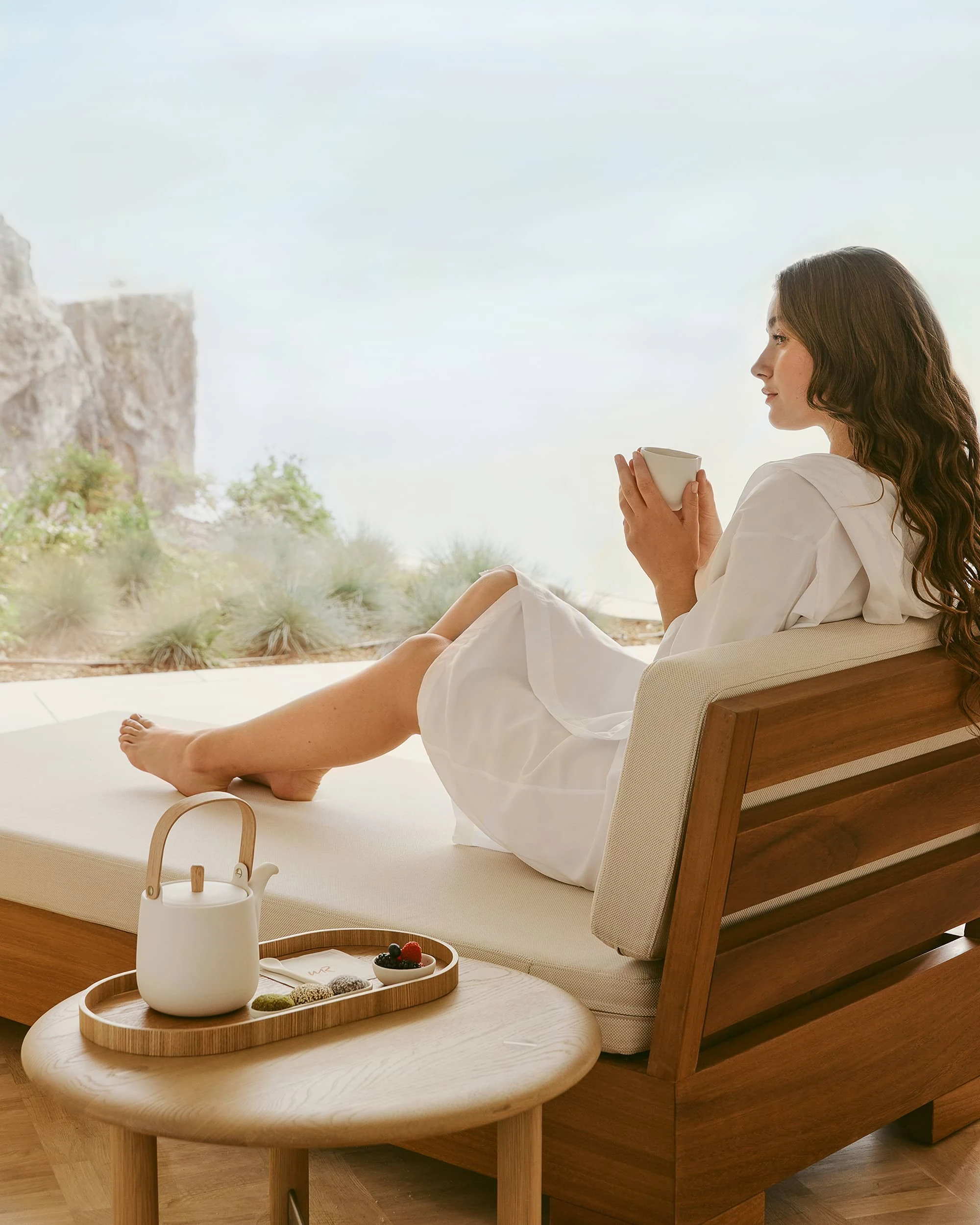 Woman in a white robe seated on a slatted timber daybed holding a cup, with a tea tray on a round oak table and the cliff visible outside.
