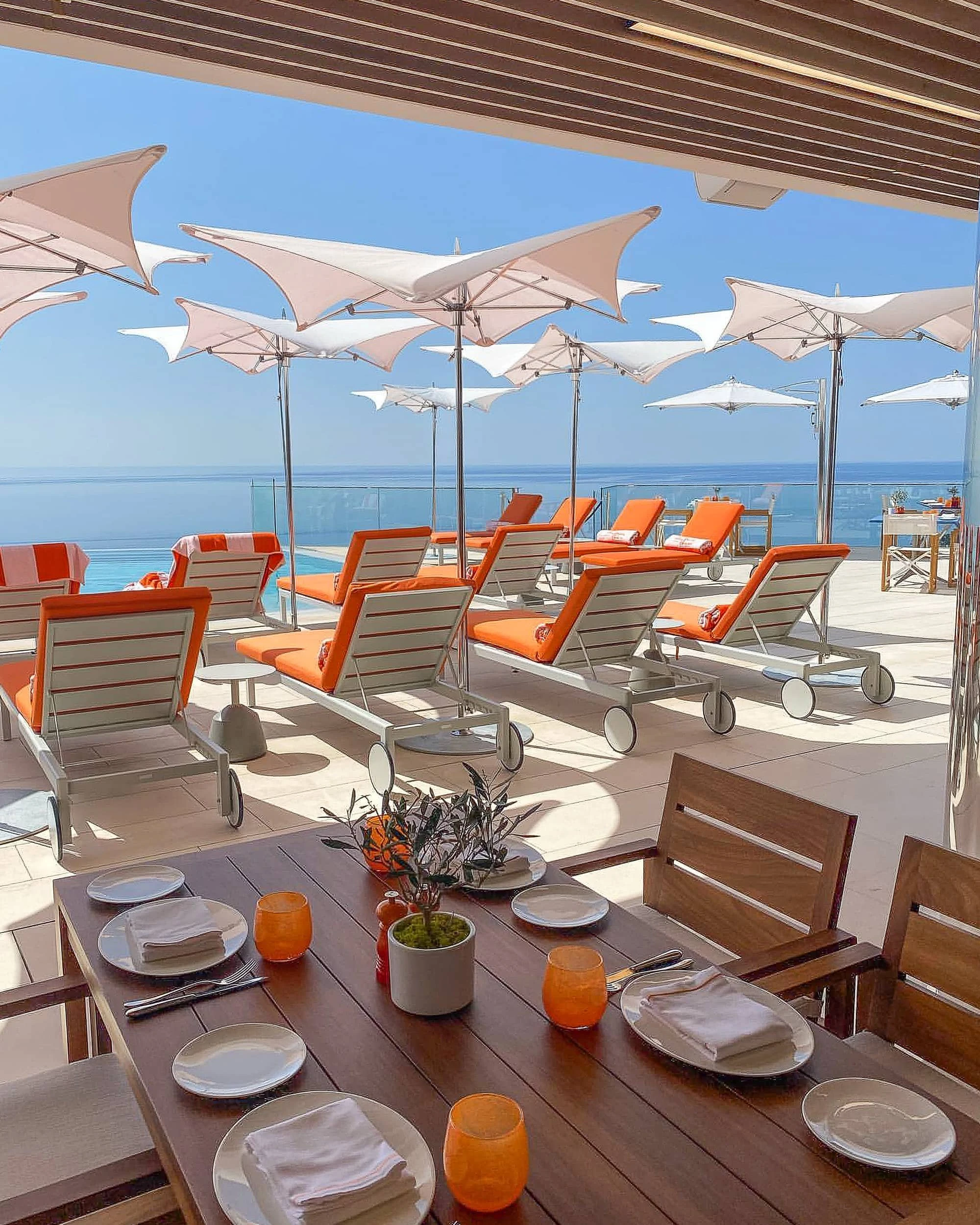 Timber table set with plates, napkins and orange glasses under a slatted overhang, with rows of orange loungers and white parasols facing the sea.