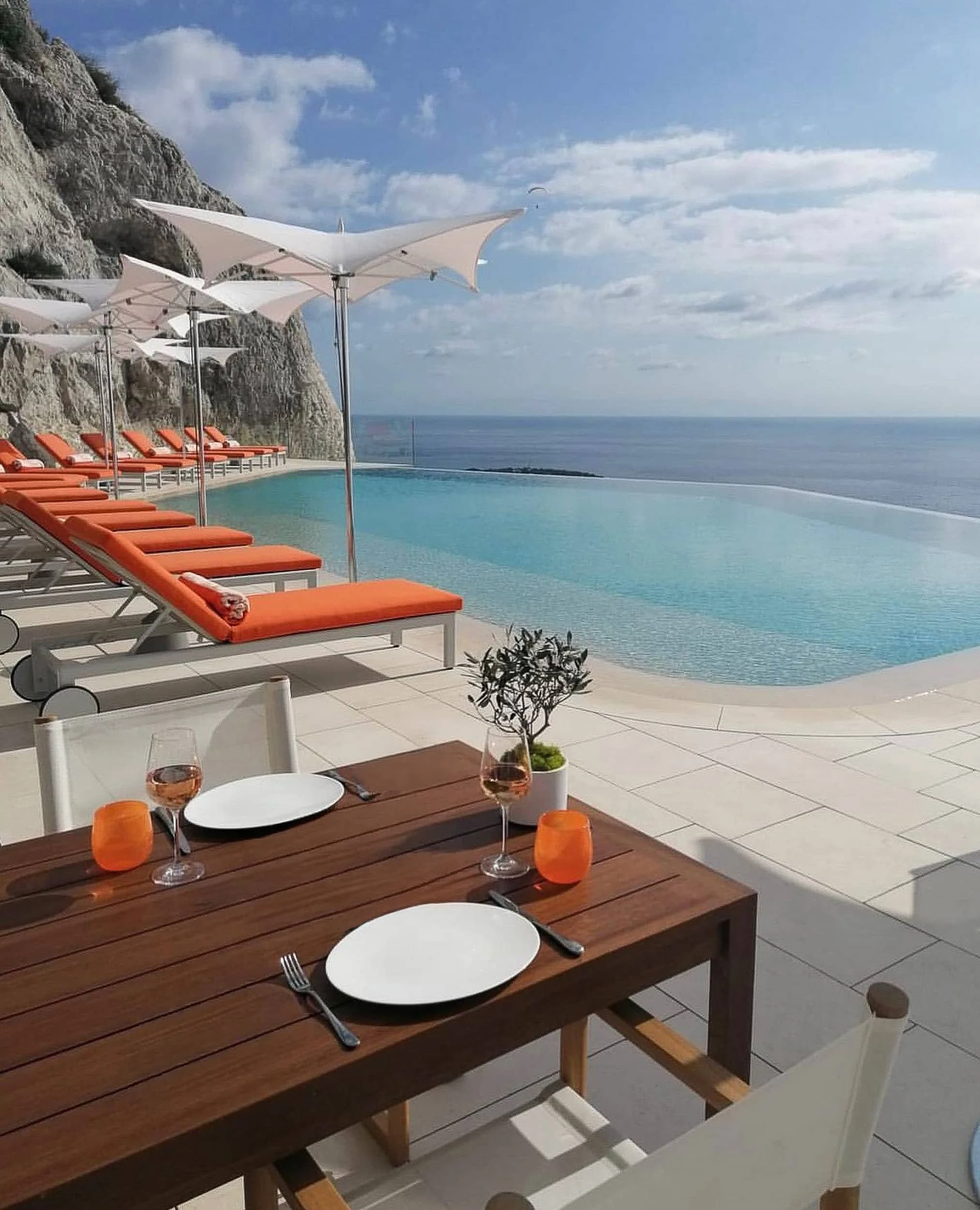 Iroko table set with plates, wine and an olive sapling beside the infinity pool, orange loungers and parasols lined against the cliff.