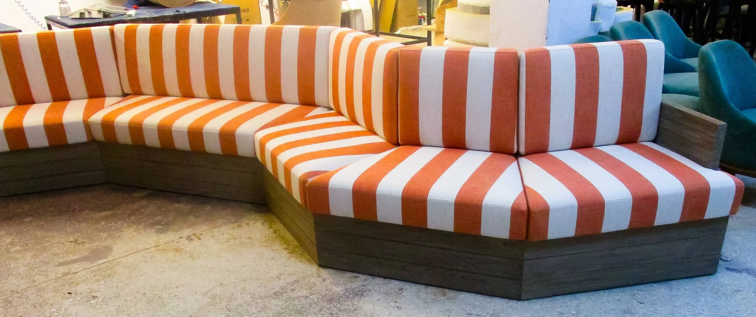 Corner section of the timber banquette with striped seat and back cushions standing on a workshop floor alongside other furniture.