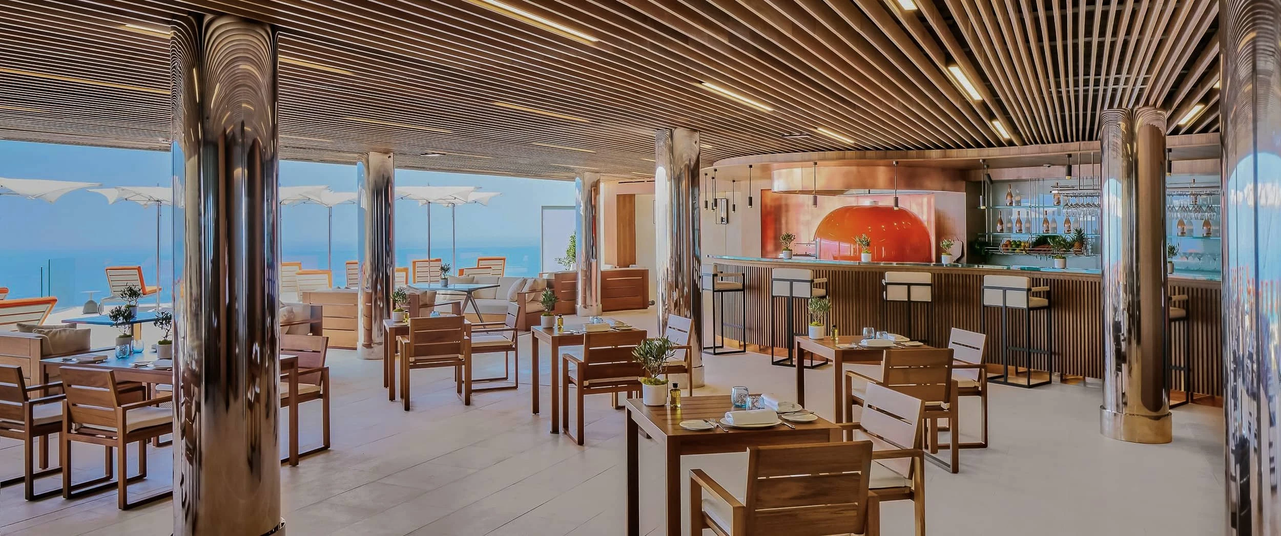 Interior of the poolside restaurant with a slatted timber ceiling, mirror-clad columns, Iroko tables and chairs and a bar with an orange pizza oven.
