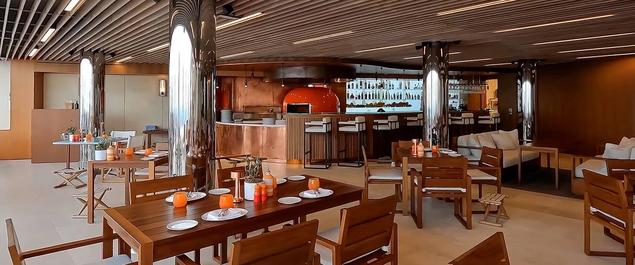 Restaurant interior at The Maybourne Riviera with timber dining tables, bar stools, an orange domed oven and a copper-clad counter under a slatted ceiling.