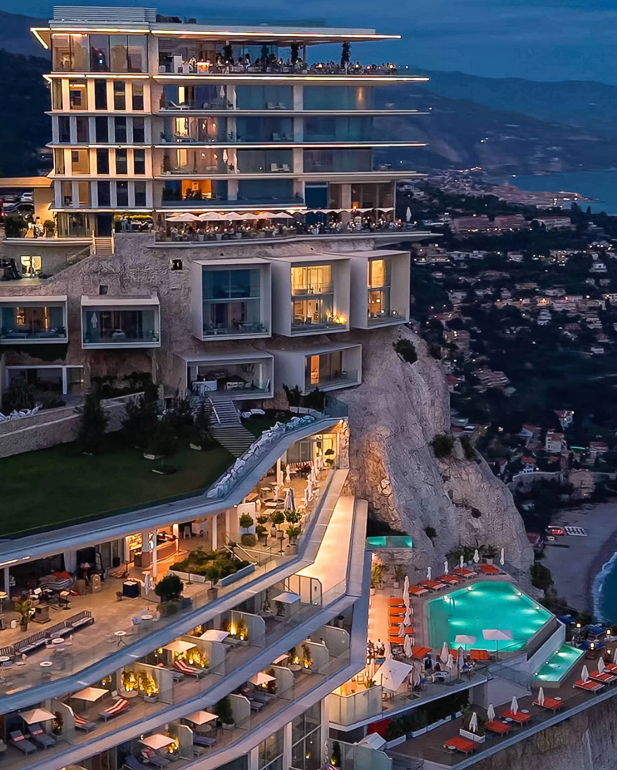 Evening aerial view of the illuminated hotel, its terraces, suite boxes and lit pool stepping down the cliff above the coast.