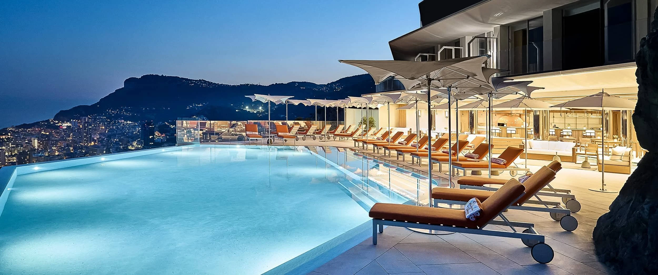 The lit infinity pool at night with orange loungers, white parasols and the open restaurant behind, Monaco's lights across the bay.