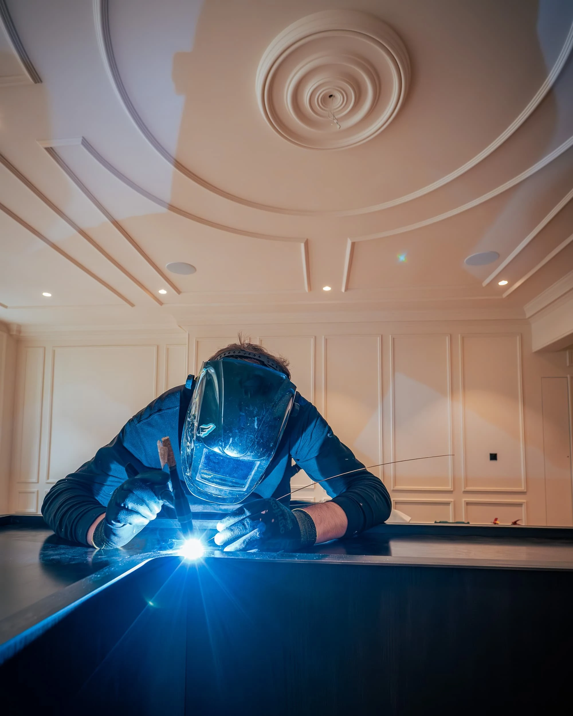 Wide view of a welder in full mask working on the blackened steel reception desk frame in the panelled lobby, arc light illuminating the ceiling and revealing the craft behind the finished desk.