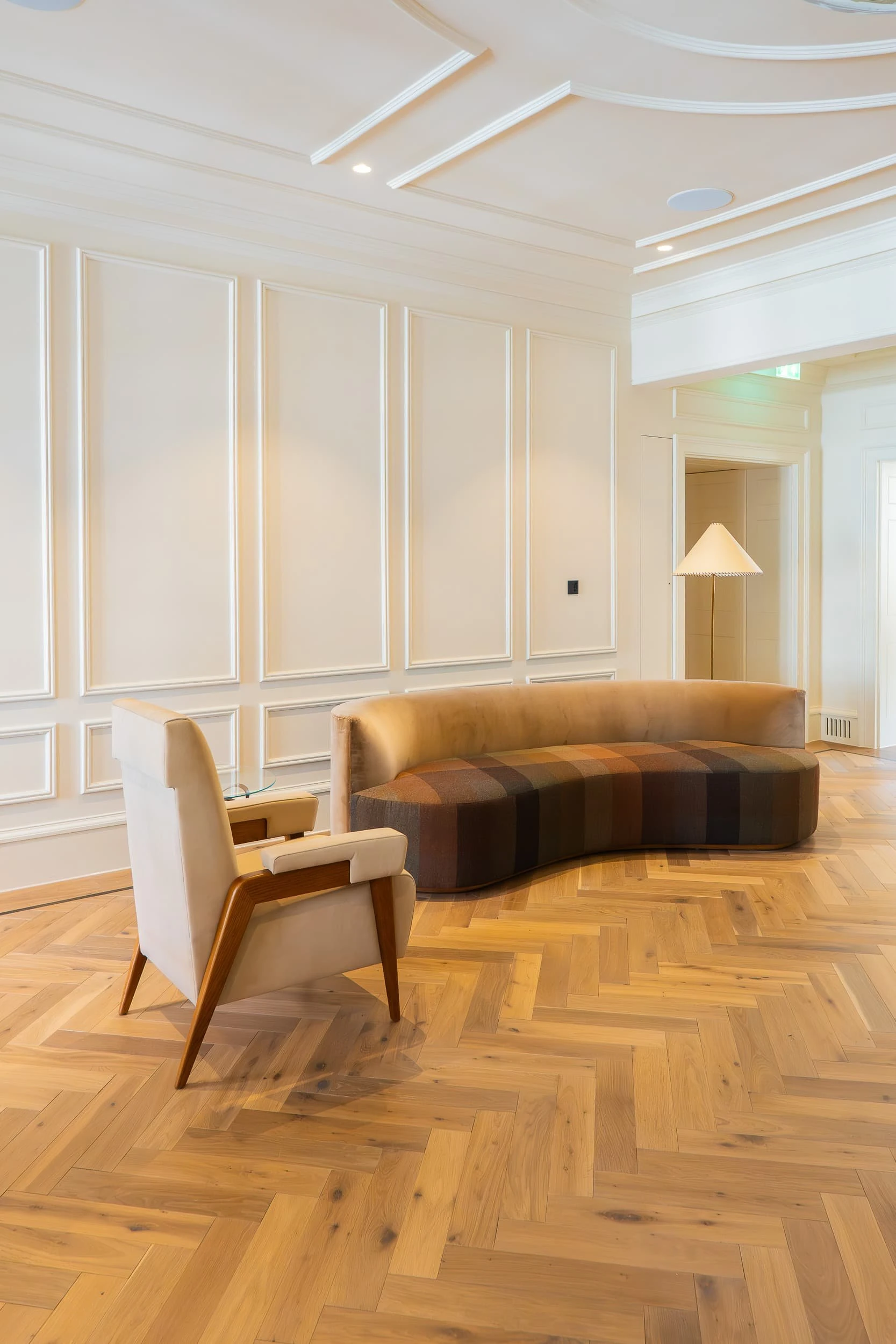 Calm corner of the lounge with a curved patchwork-upholstered sofa, mid-century armchair and floor lamp against cream panelling and herringbone oak floor, bringing a domestic mood to the office.