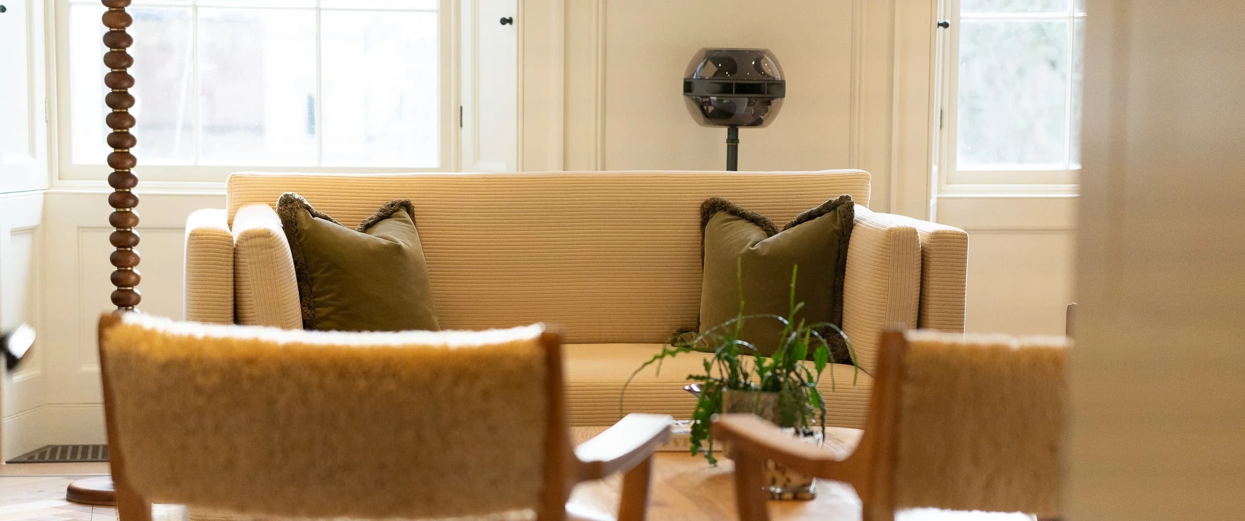 Framed view towards a cream sofa with green cushions and shearling chairs in the foreground, with a sculptural black speaker behind, capturing the quiet, domestic feel of the meeting area.