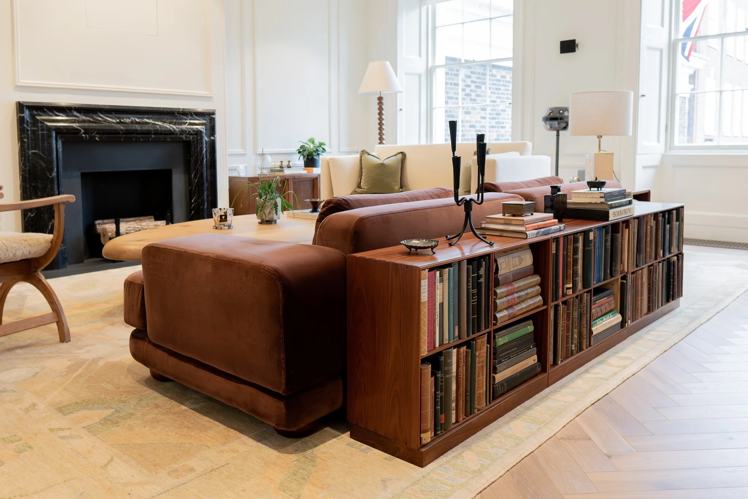 Lounge arrangement with brown velvet sofa backed by a long low bookcase of vintage volumes, cream sofa and black marble fireplace, giving the office the relaxed feel of a cultured city apartment.