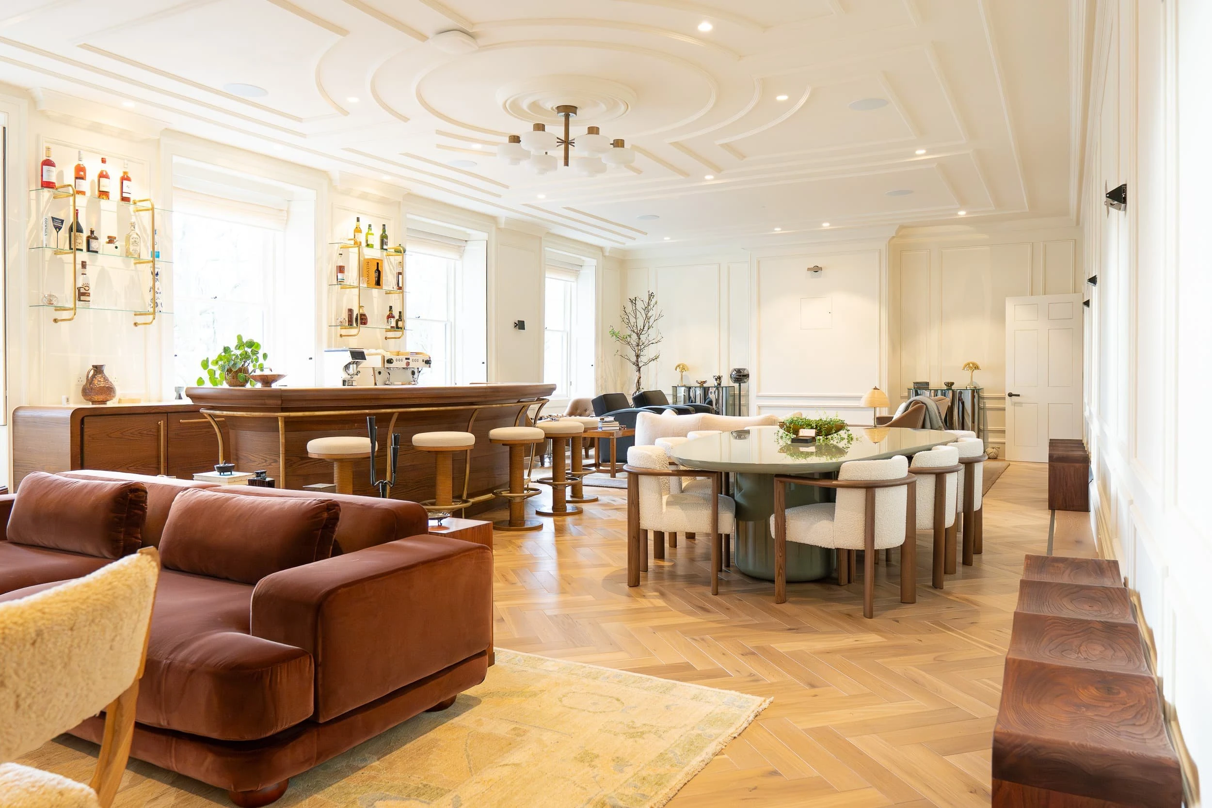 View across the bespoke bar run, showing oak cabinetry, brass footrail and upholstered stools, drawing attention to the crafted joinery and soft finishes within the office's social space.