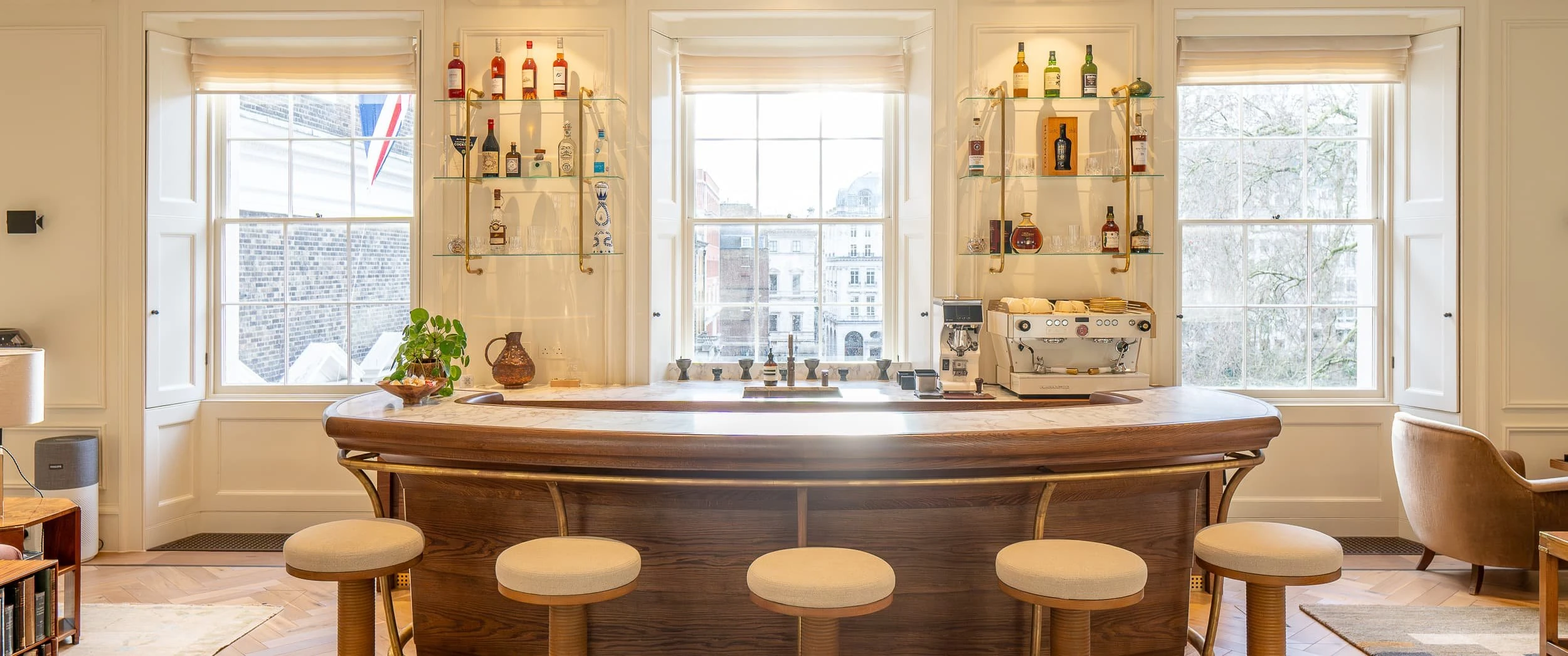 Wide perspective of the bar and dining area in the main room, with curved timber bar, upholstered stools, round dining table and brown velvet sofa, creating a hospitality-style hub within the office.