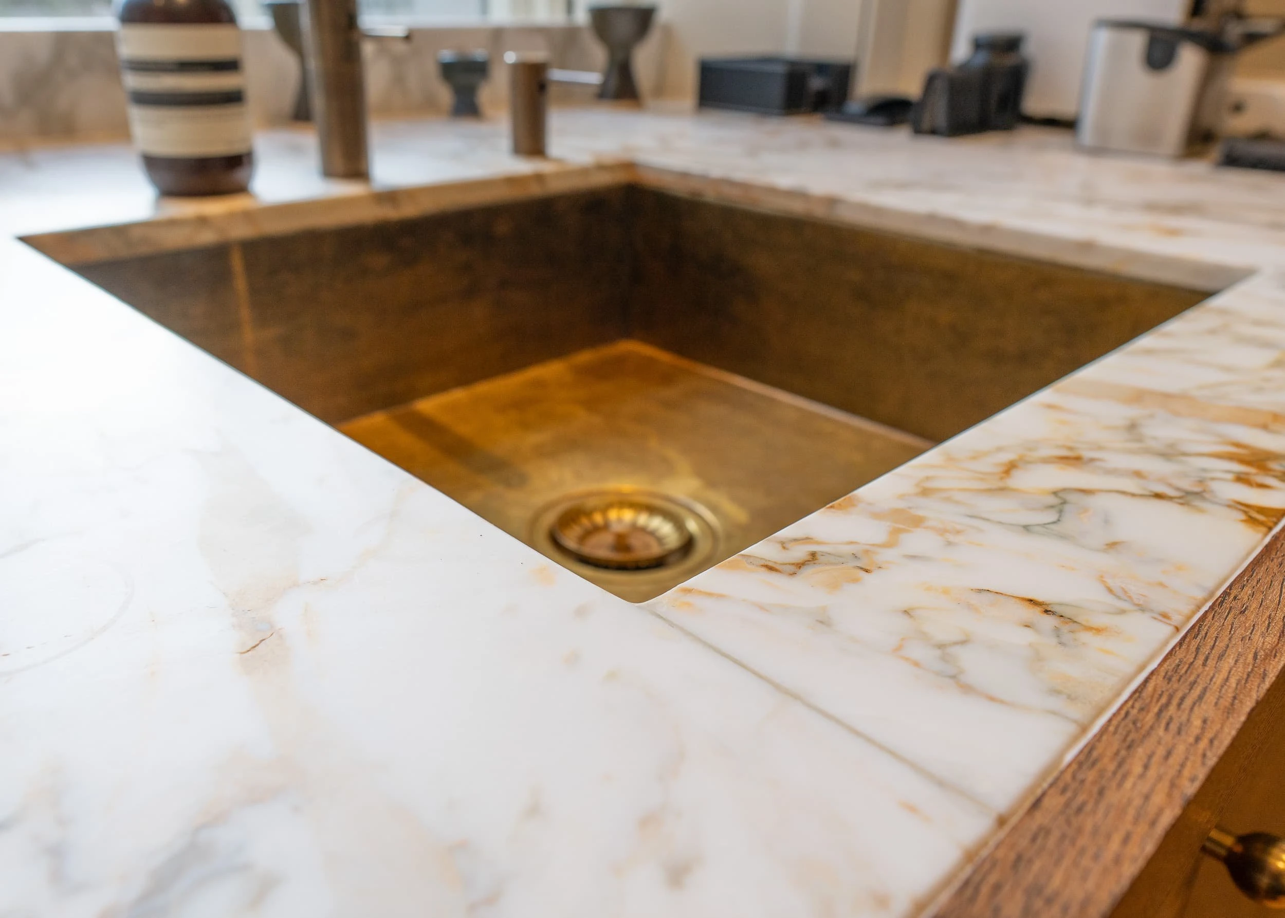 Close-up of a polished brass sink set into a veined marble worktop, with brushed taps and oak joinery below, illustrating the level of detail given to the bar and pantry areas in the office.