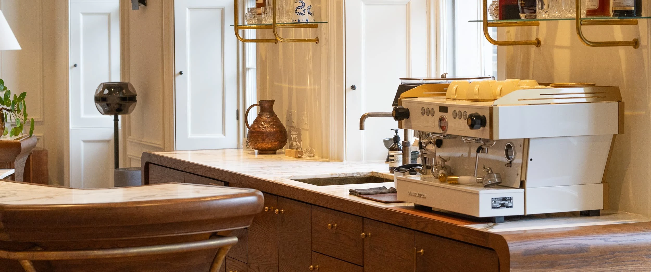 Marble-topped bar counter with espresso machine, brass-framed shelves and oak cabinetry beneath, reinforcing the blend of members' club comfort and functional office refreshment point.