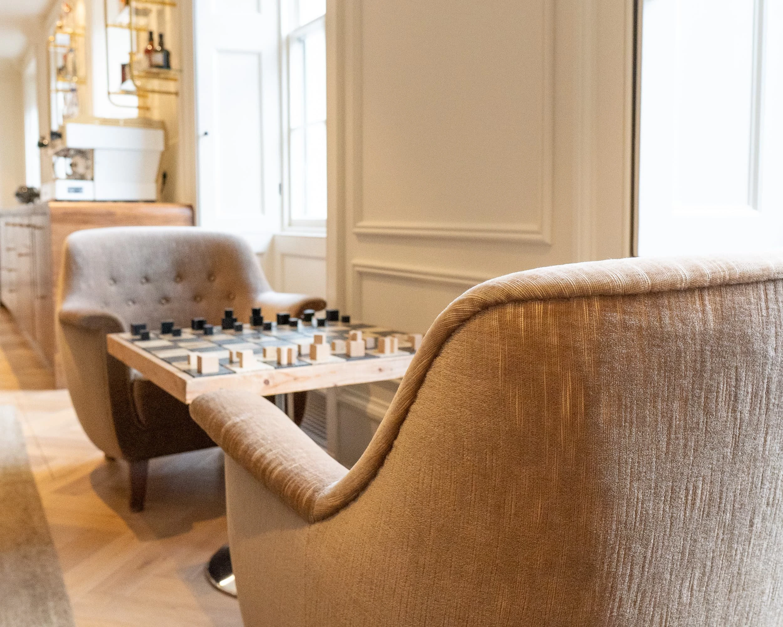 Pair of tufted lounge chairs upholstered in soft neutral fabric facing a chessboard table, with bar and storage beyond, creating an inviting corner for informal breaks within the offices.