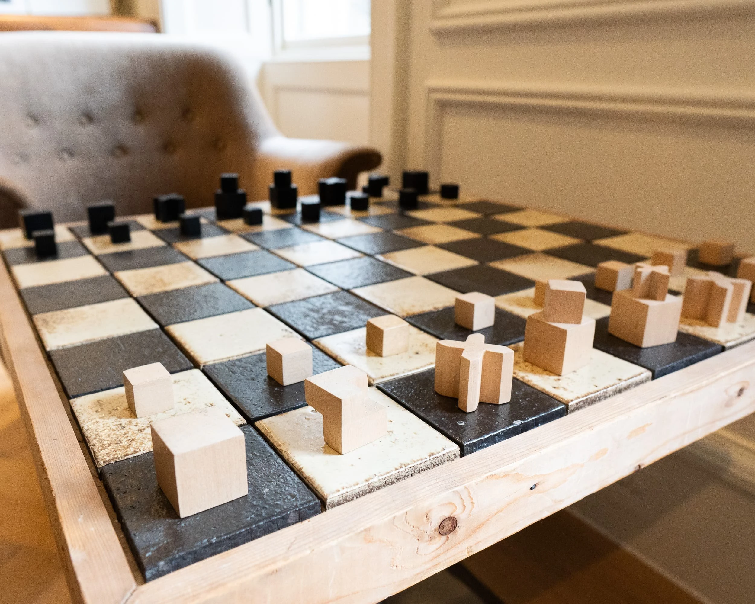 Handmade chessboard table with chunky wooden pieces on glazed tiles, set before a tufted lounge chair, providing a playful, club-like detail within the relaxed lounge of the private offices.