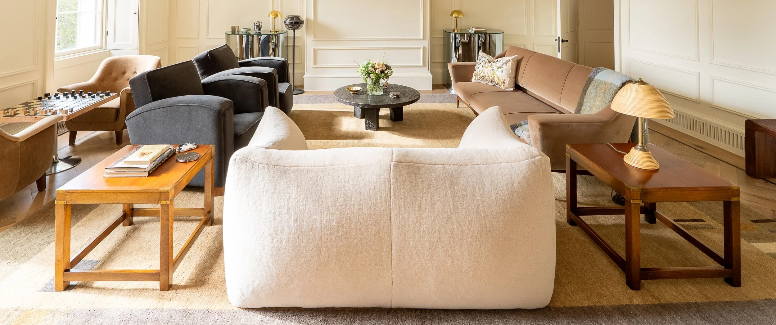 Wide shot across the principal lounge from behind a soft ivory sofa, revealing rows of armchairs, coffee tables and a long tan sofa, making the private office feel like a generous city living room.