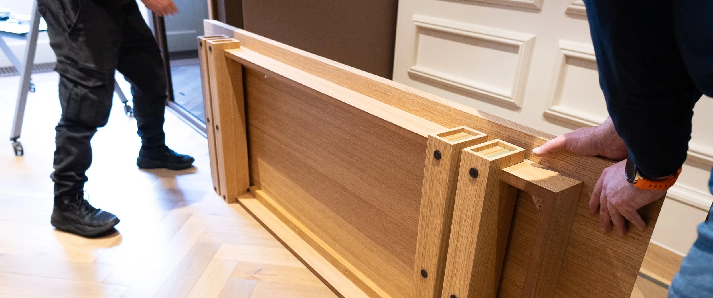 Installation shot of two fitters carrying a substantial oak table base across herringbone flooring, showing the weight and care involved when positioning heavy bespoke elements in the office.