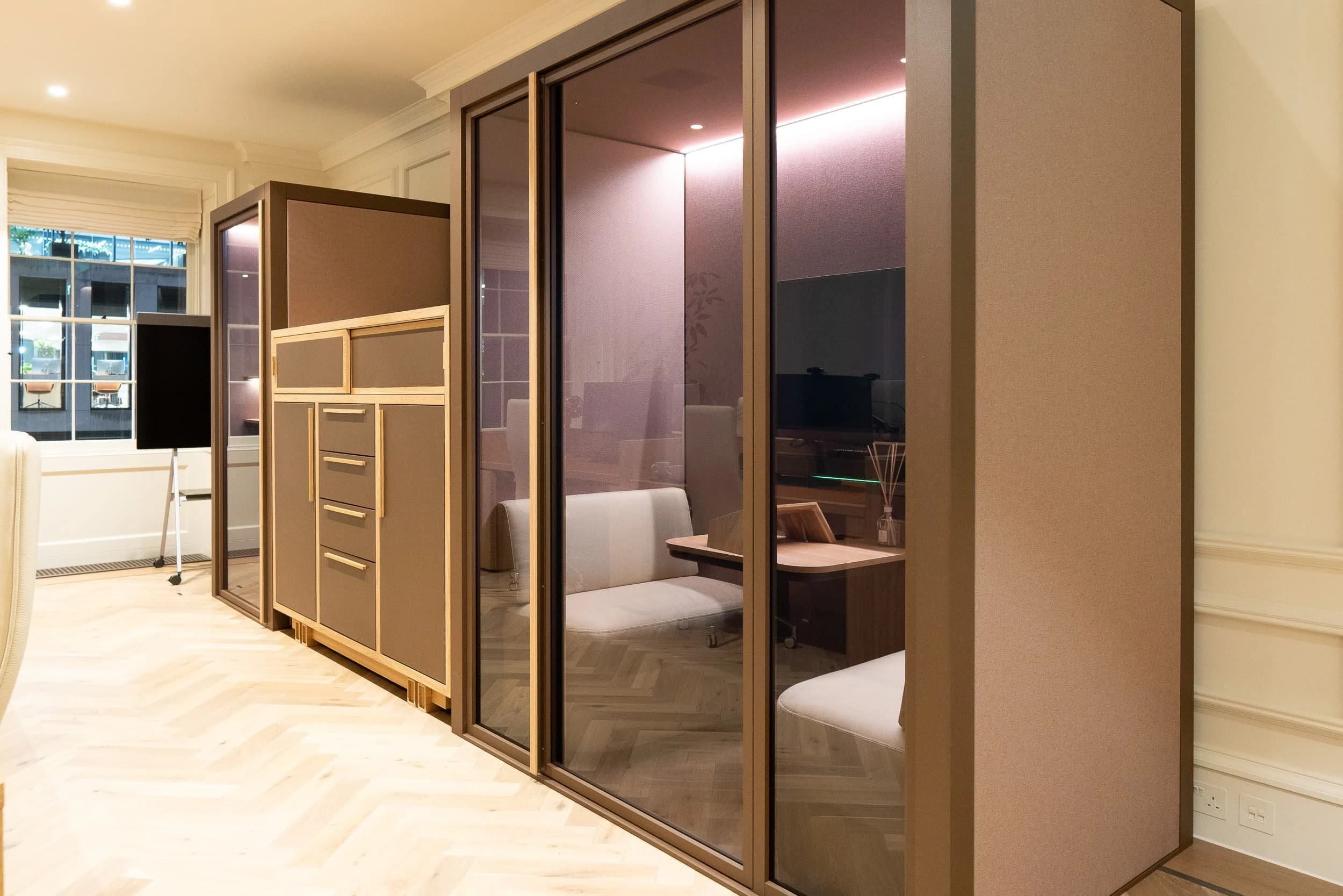 Night-time shot of taupe and oak storage unit framed between glazed partitions, seen from the reception desk, with a small plant in the foreground, underlining the calm, ordered feel of the workspace.
