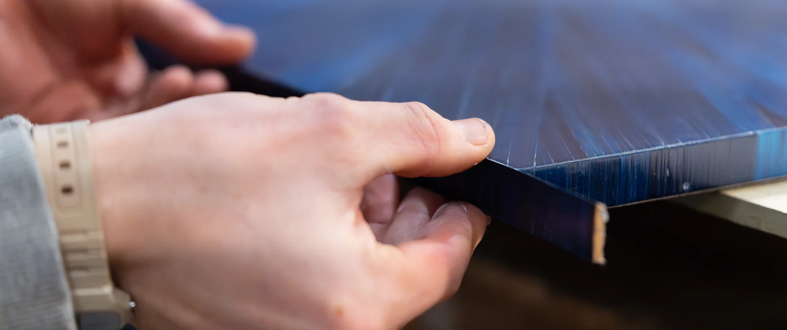Maker running a fingertip along the edge of a finished blue straw-marquetry panel to check the lay of the fibres.