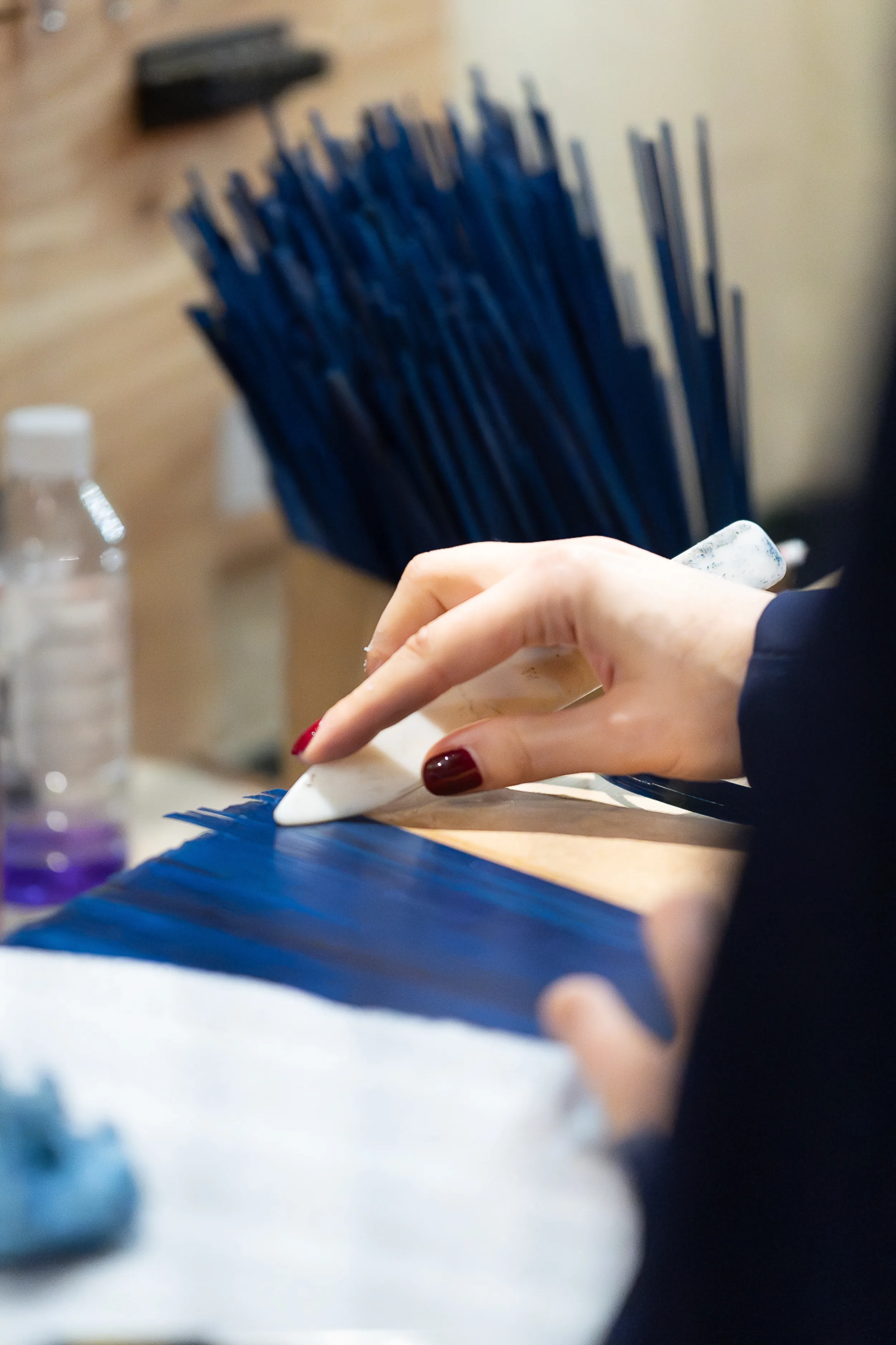 A maker combing dyed straw into place with a bone folder, a bundle of blue straw standing at the bench behind.