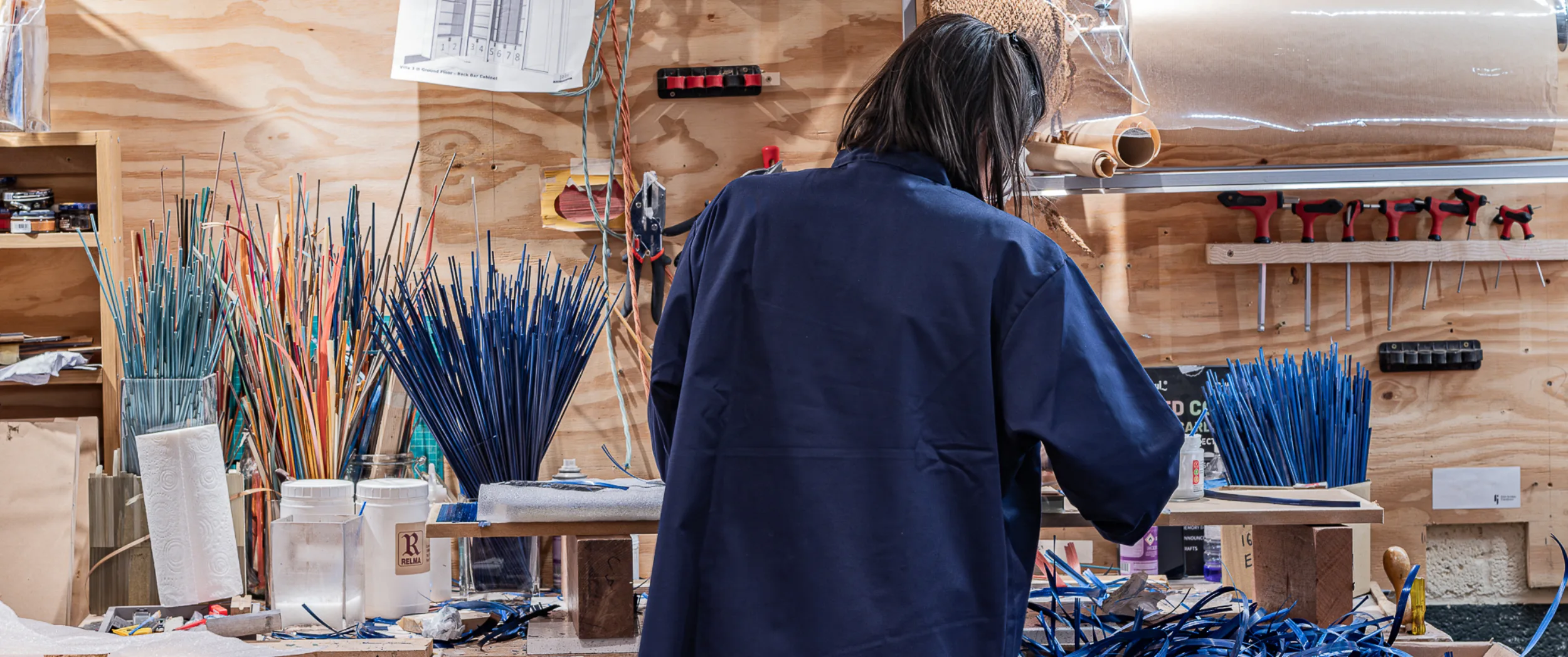 A marqueteur at the bench in the East London workshop, bundles of dyed straw standing in jars and the door schedule pinned to the wall above.