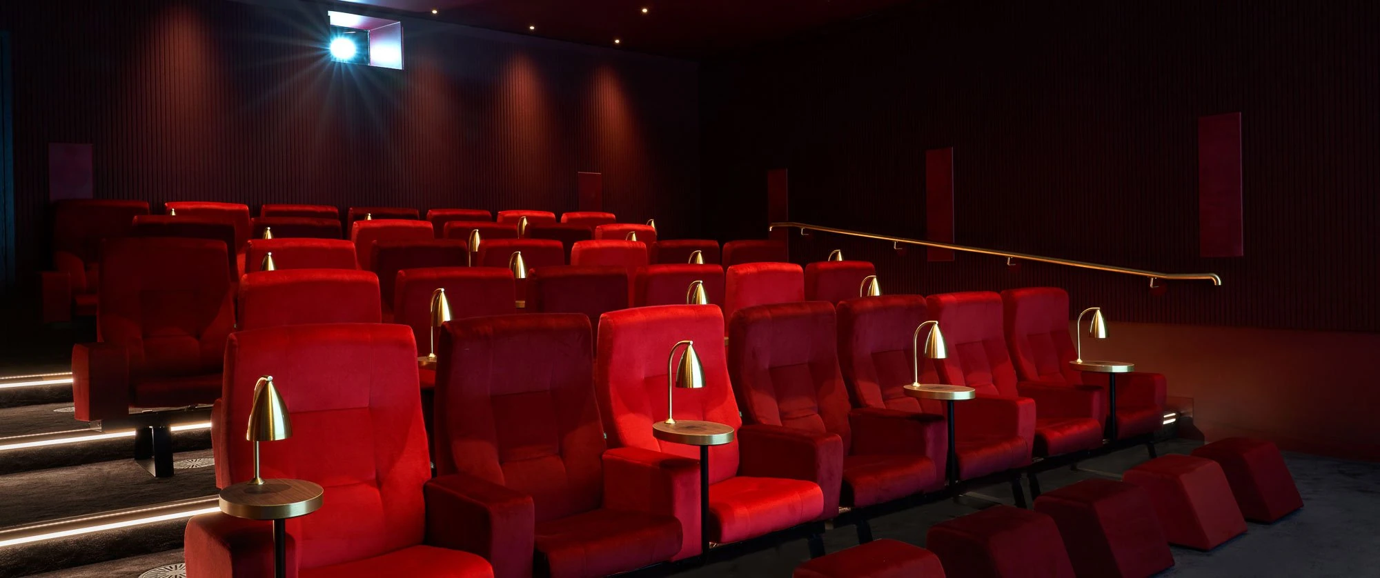 Rows of red velvet recliners with brass side tables and reading lamps in the private screening room at The BoTree, with a projector beam above the ribbed dark walls.