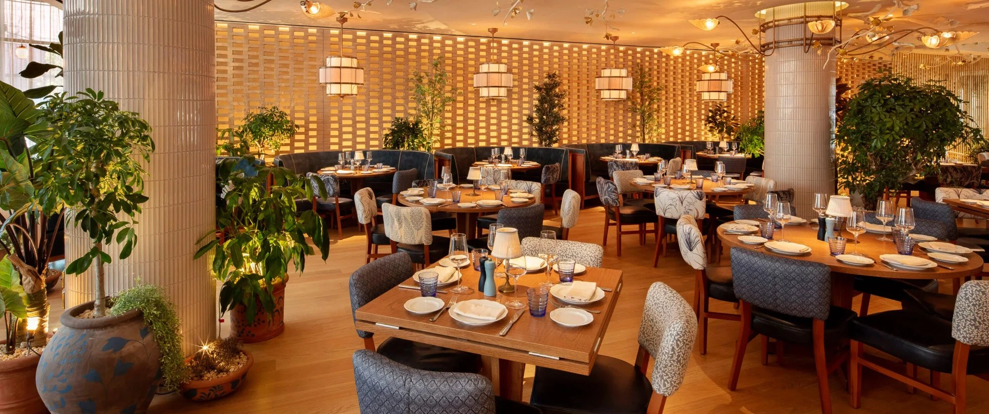 Dining tables and patterned chairs at LAVO London set before a backlit woven brick screen, with dark blue fitted banquettes and potted plants along the room.