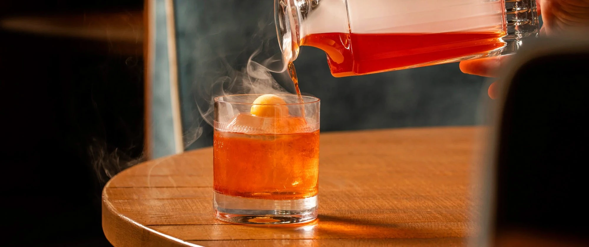 A cocktail poured from a glass carafe into a tumbler on a timber table at LAVO London, with smoke drifting across the frame.