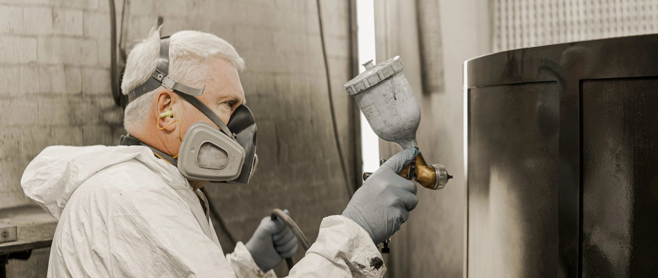 Close view of a Make Bespoke Studio finisher in respirator and gloves holding a gravity-fed spray gun beside a black-lacquered banquette carcass in the workshop booth.