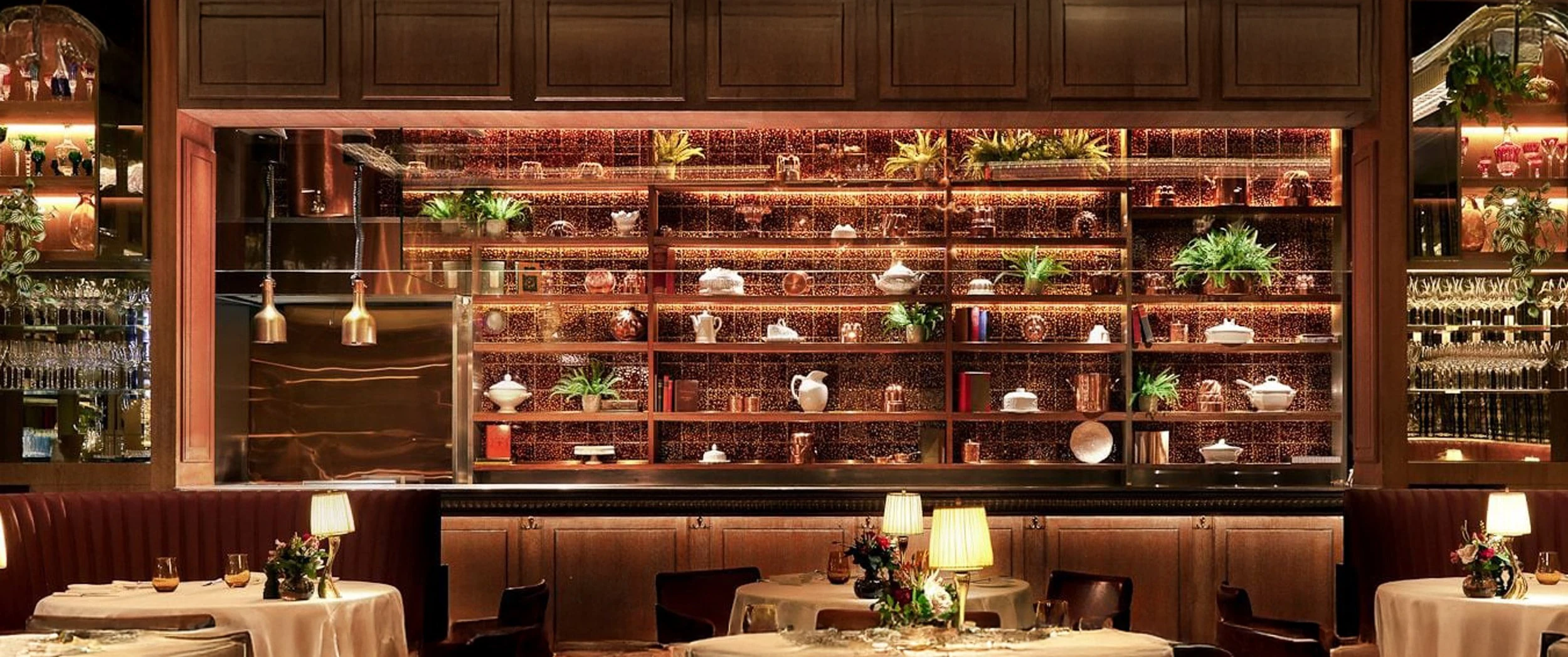 Interior of The Grill at The Dorchester showing the lit copper shelving above the pass, panelled walls, fluted leather banquette seating and laid tables.