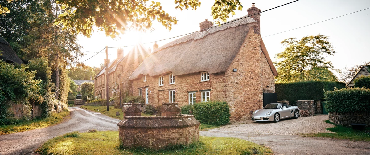 Thatched ironstone cottage at Well Cottage, Ledwell, seen from the village lane with a stone well head, clipped hedging and gravel parking in front.