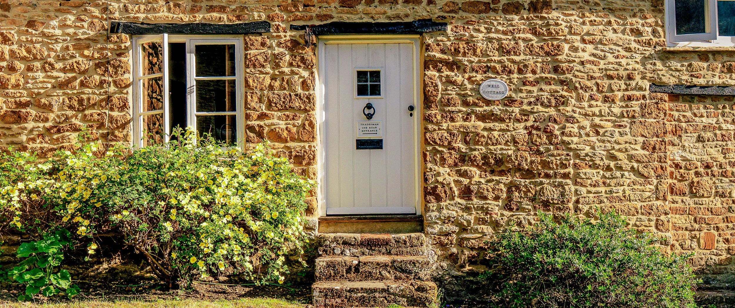 Painted timber front door with iron knocker and oval Well Cottage name plaque set into the rubble ironstone facade in Ledwell, with a yellow rose beside the casement window.